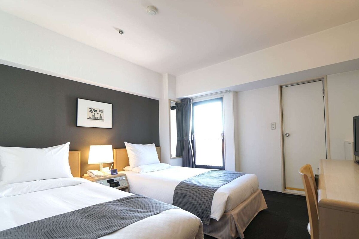 Ein Hotelzimmer mit zwei Betten, einem Schreibtisch, einem Stuhl, einem Fernseher und einem Fenster mit Vorhängen.