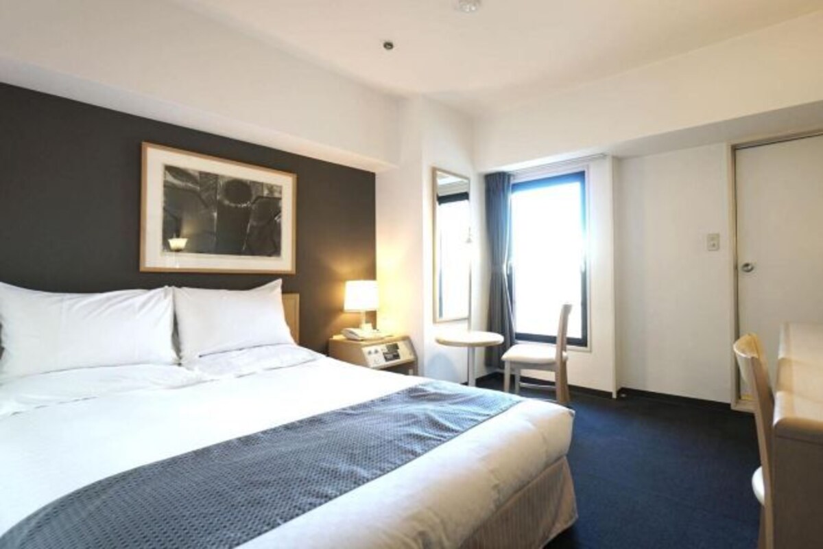 Una habitación de hotel con cama, mesita de noche con lámpara, silla, ventana y puerta.