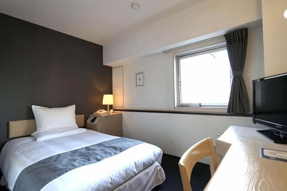 Une chambre d’hôtel comprenant un lit, un bureau, une télévision, une fenêtre avec des rideaux et une table de chevet avec une lampe.