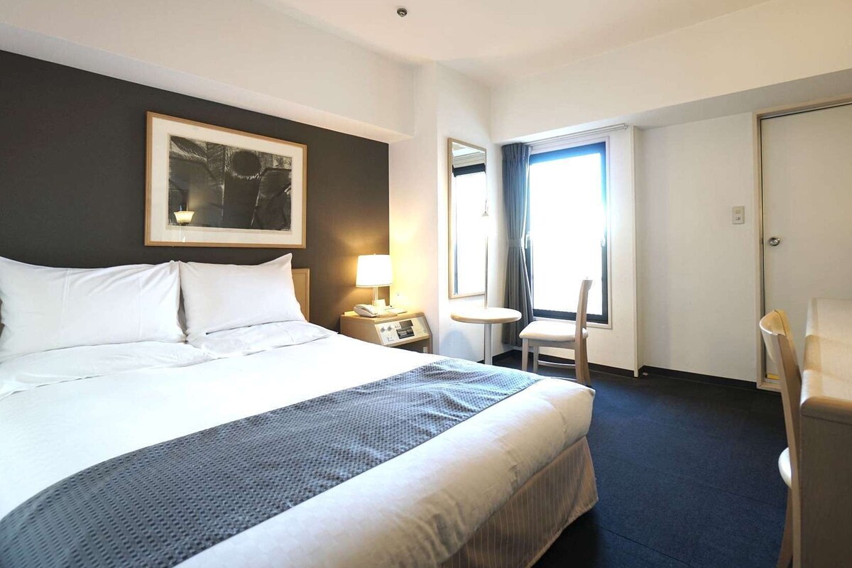 Ein Hotelzimmer mit einem Bett, einem Nachttisch mit Lampe, einem Sessel, einem kleinen Tisch und einem Fenster mit Vorhängen.