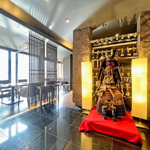 A modern interior with a traditional Japanese warrior statue on a red cloth, a bar area with bottles, and a seating area with tables and chairs. - HOTEL THE CENTRE UTSUNOMIYA (Utsunomiya)