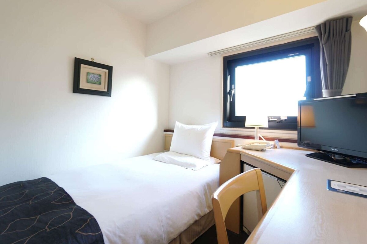 Ein Hotelzimmer mit einem Bett, einem Schreibtisch mit Computer, einem Sessel, einem Fernseher und einem Fenster mit Vorhängen.