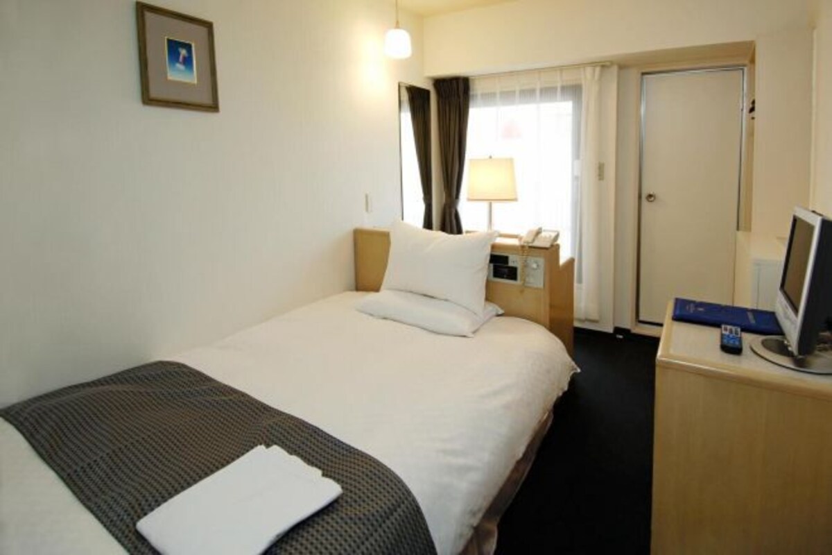 Habitación de hotel con cama, escritorio, computadora, teléfono y ventana con cortinas.