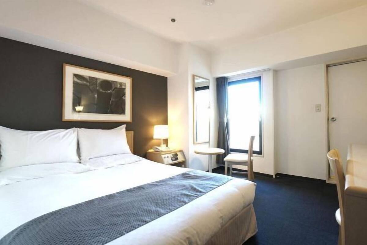 Une chambre d’hôtel avec un lit, une table de chevet avec une lampe, une chaise, une fenêtre et une porte.