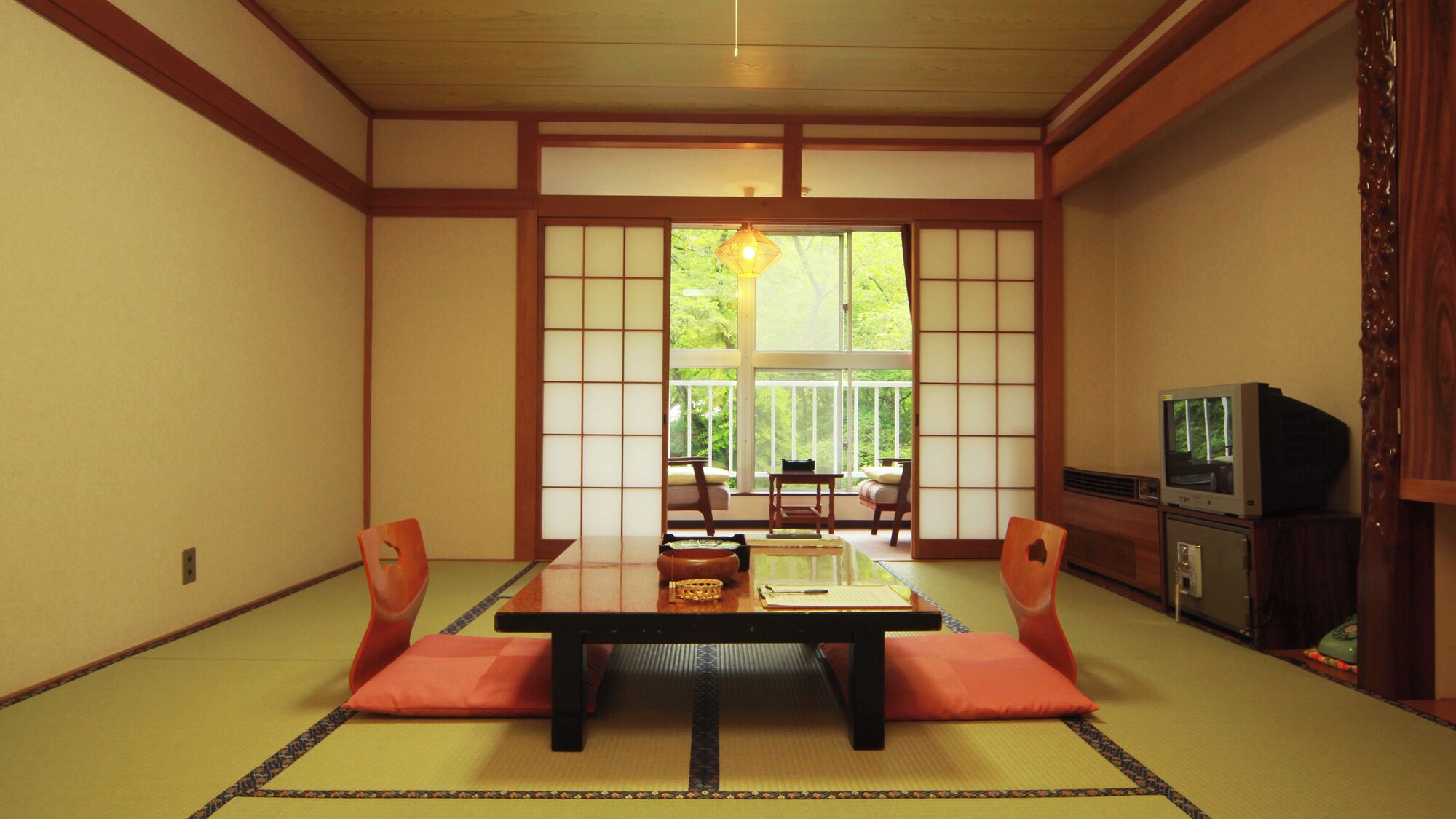 A traditional Japanese room with tatami flooring, a low table with cushions, a television, and a sliding door leading to a balcony.