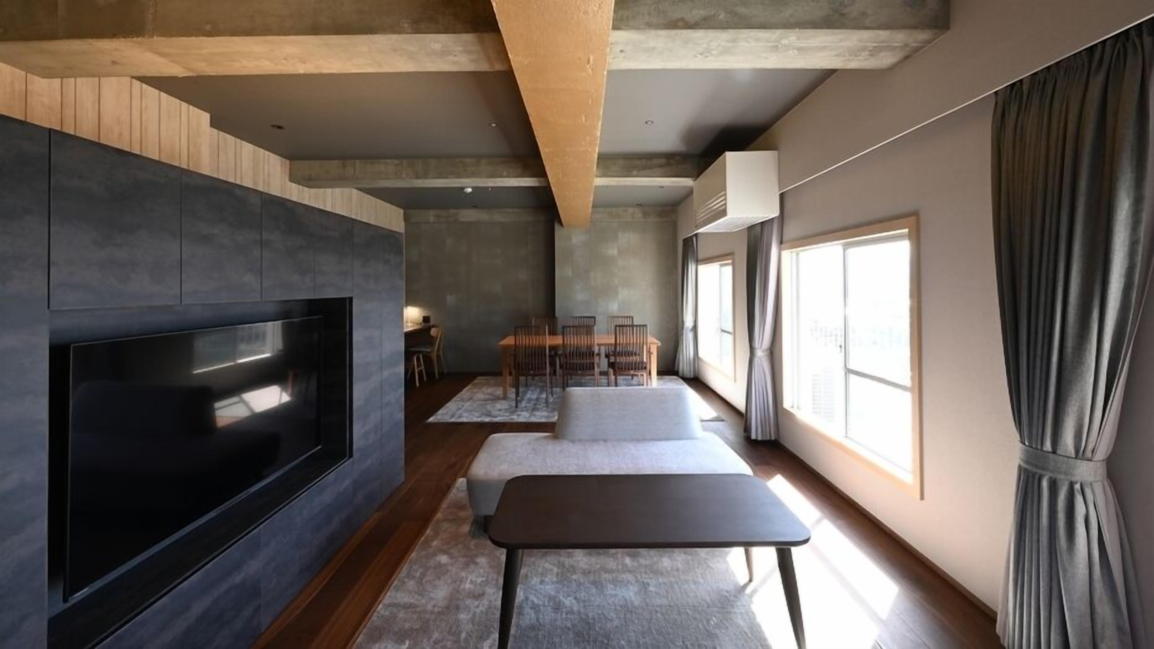 A modern living room with a flat-screen TV, a sofa, a dining table with chairs, and large windows.