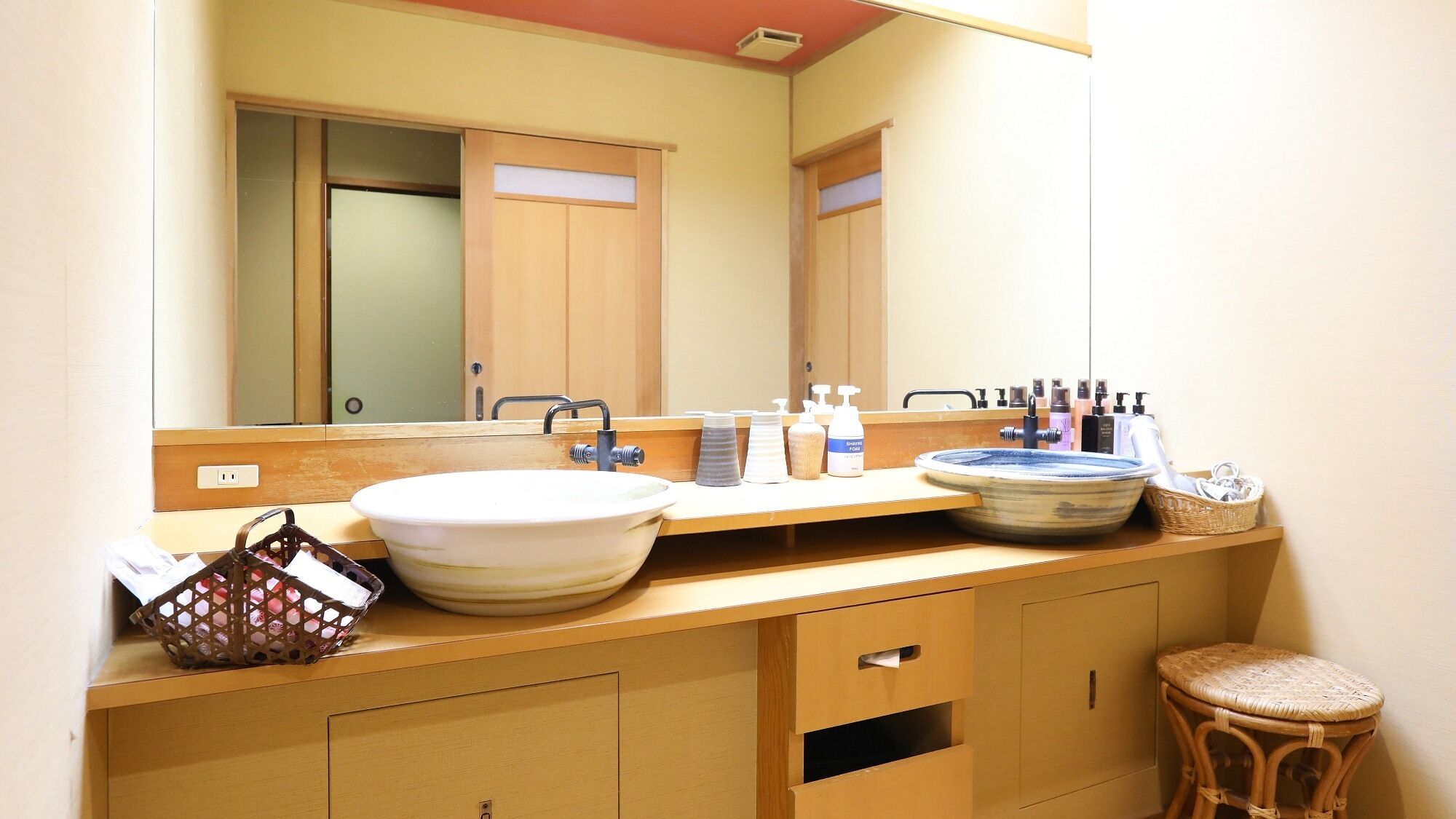 A bathroom with a large mirror, wooden vanity, a bowl sink, and a wicker stool.