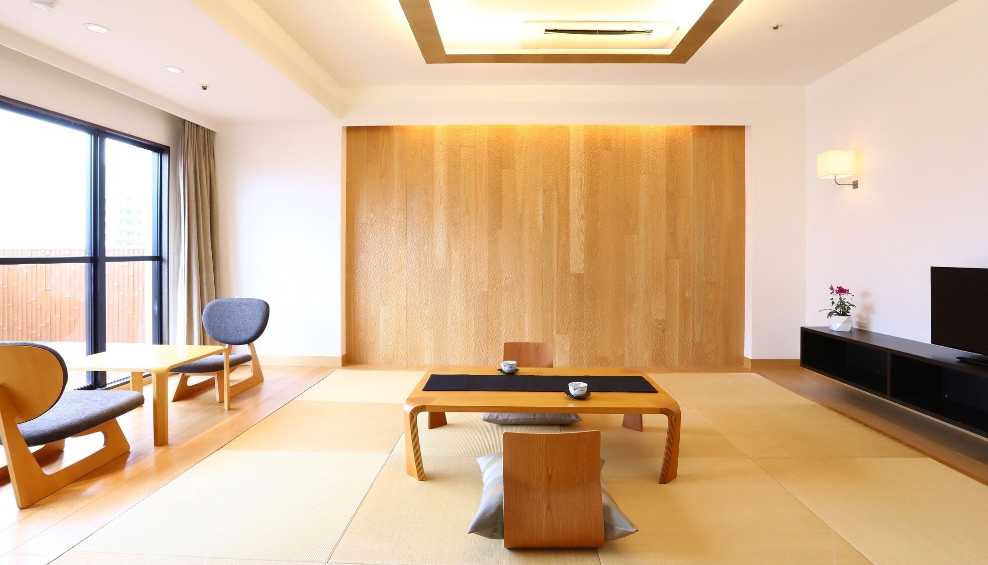 A modern hotel room with a large window, a dining table, two chairs, a sofa, a TV, and a wall-mounted shelf.