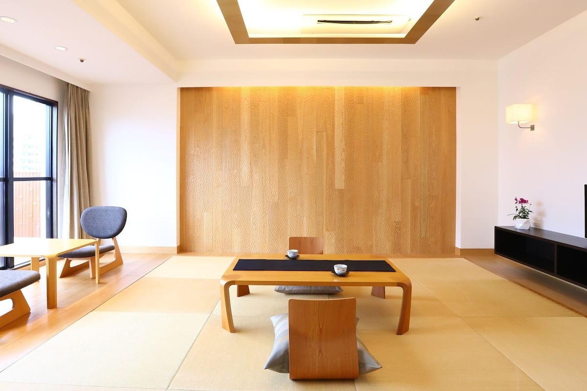 A modern hotel room with a large window, a dining table, two chairs, a sofa, a TV, and a wall-mounted shelf.