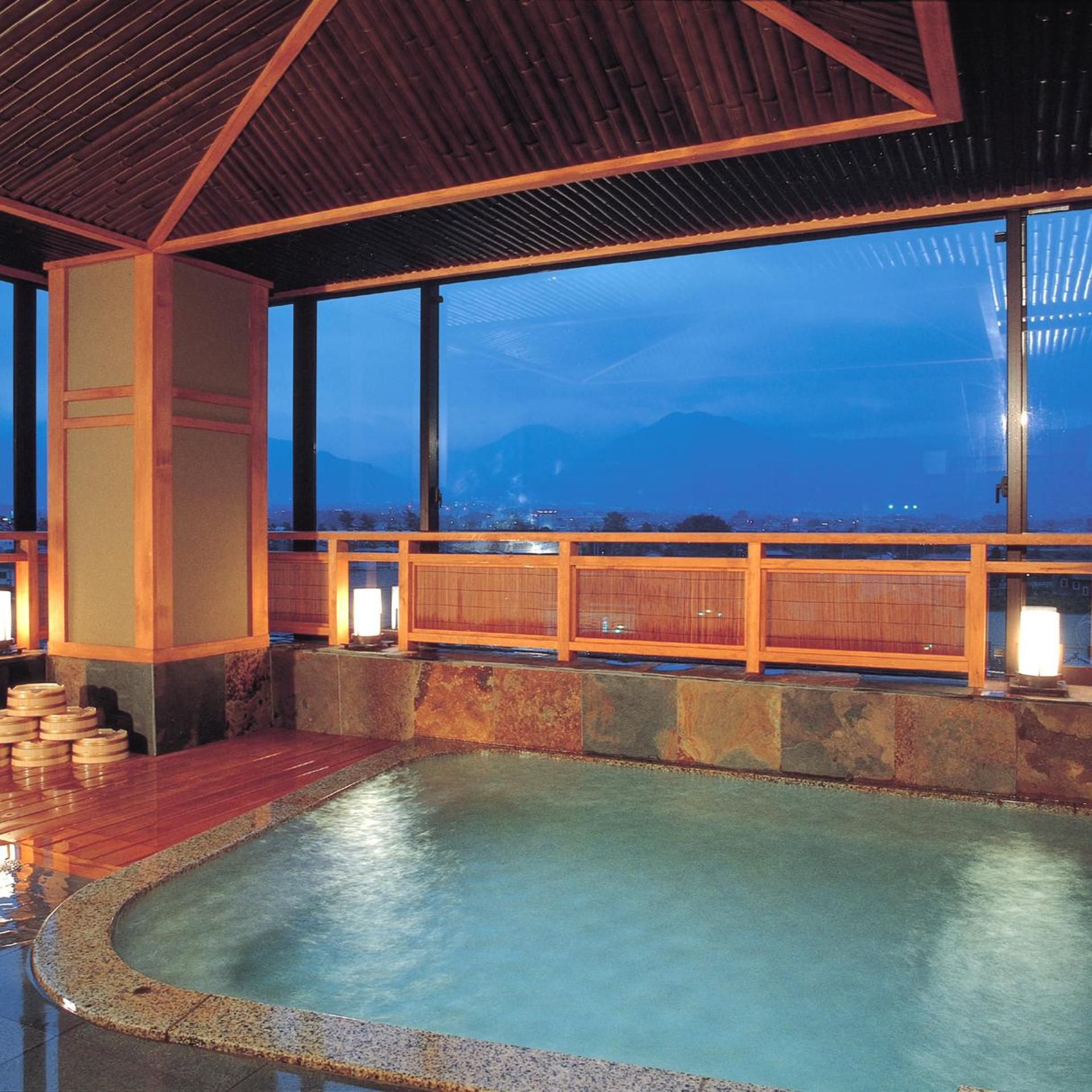 A traditional Japanese onsen with a wooden interior, a stone bath, and a view of the cityscape and mountains at dusk.