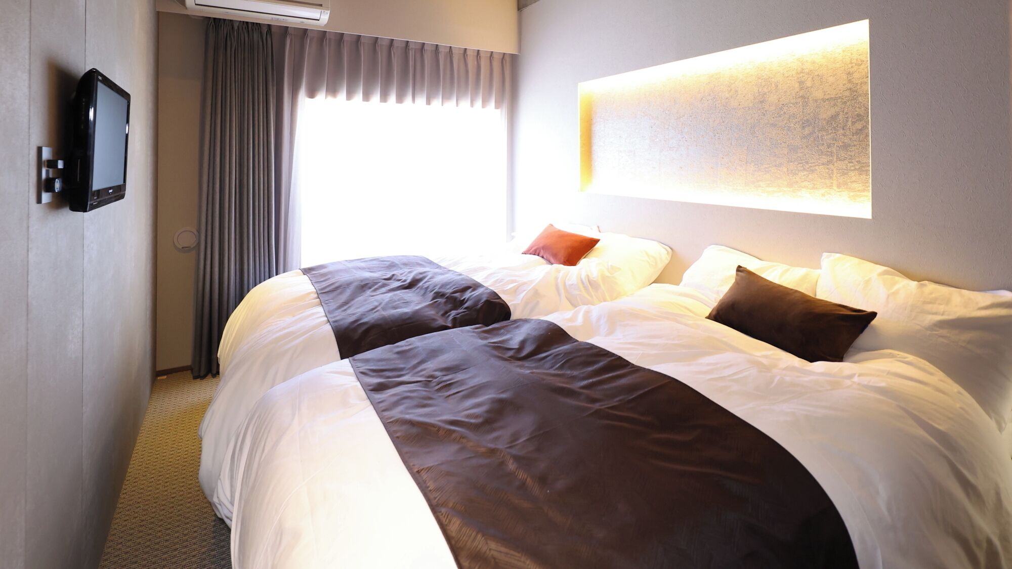 A hotel room with two beds, a flat-screen TV, a large window with curtains, and a wall-mounted light fixture.