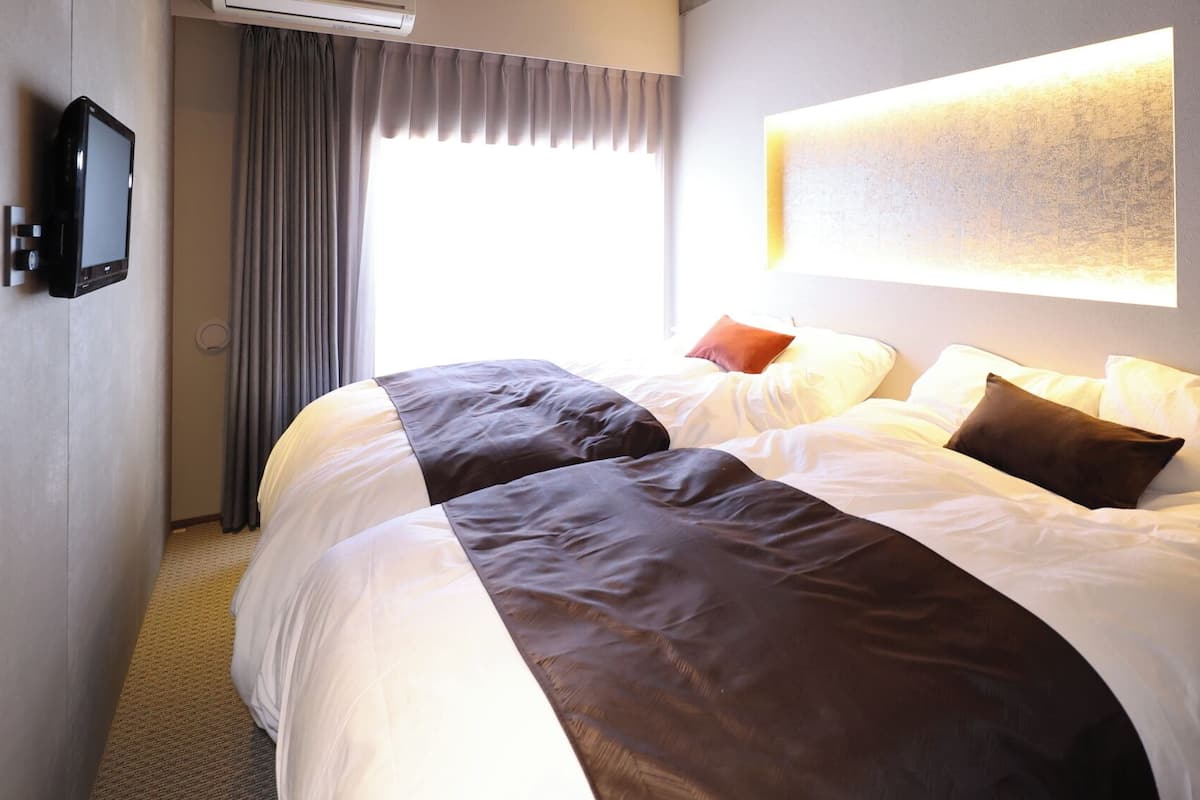 A hotel room with two beds, a flat-screen TV, a large window with curtains, and a wall-mounted light fixture.