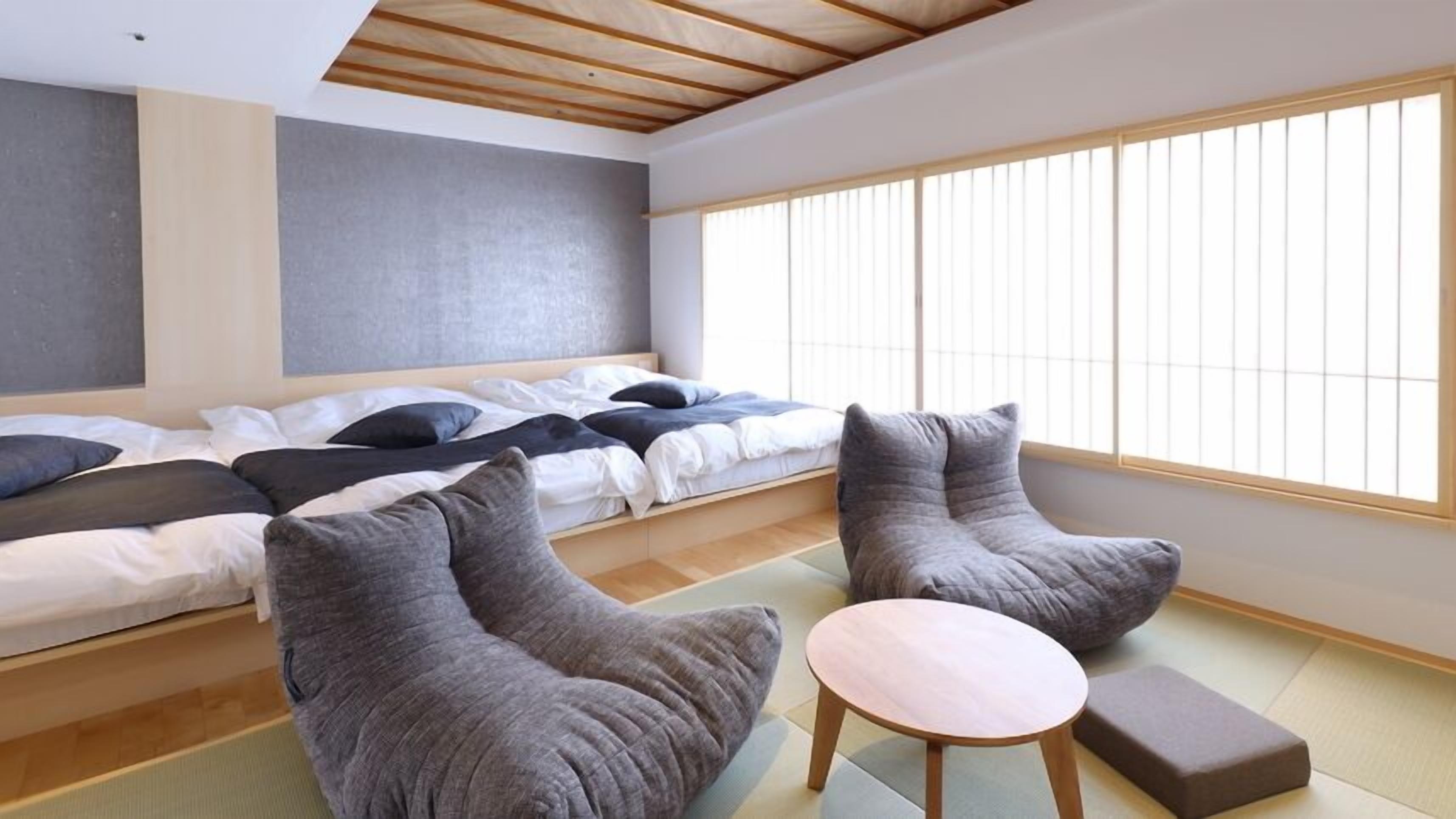 A room with a bed, two armchairs, a small table, and a large window with blinds.