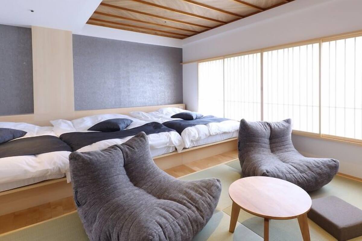 A room with a bed, two armchairs, a small table, and a large window with blinds.