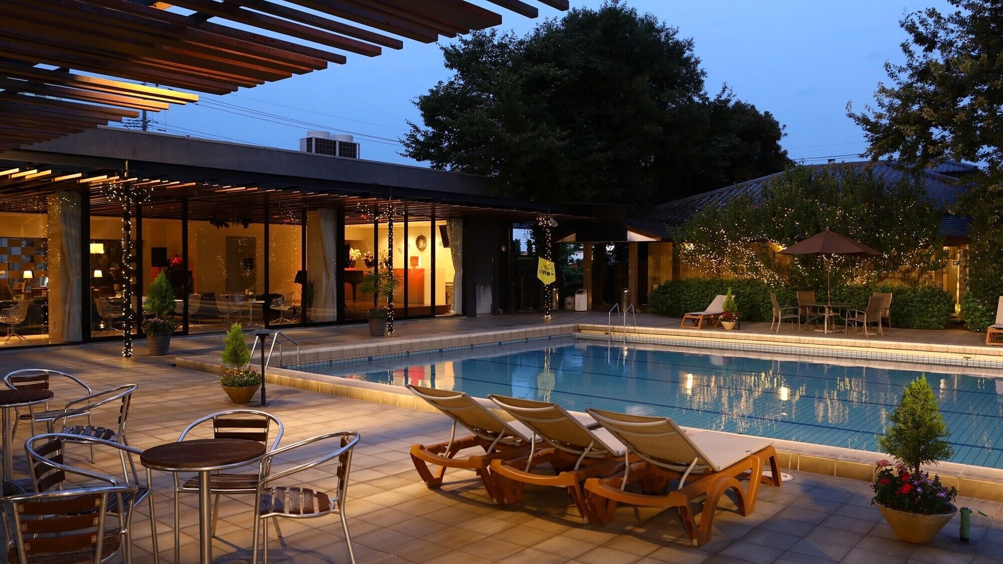 A poolside area with lounge chairs, a dining table, and a restaurant with glass doors.