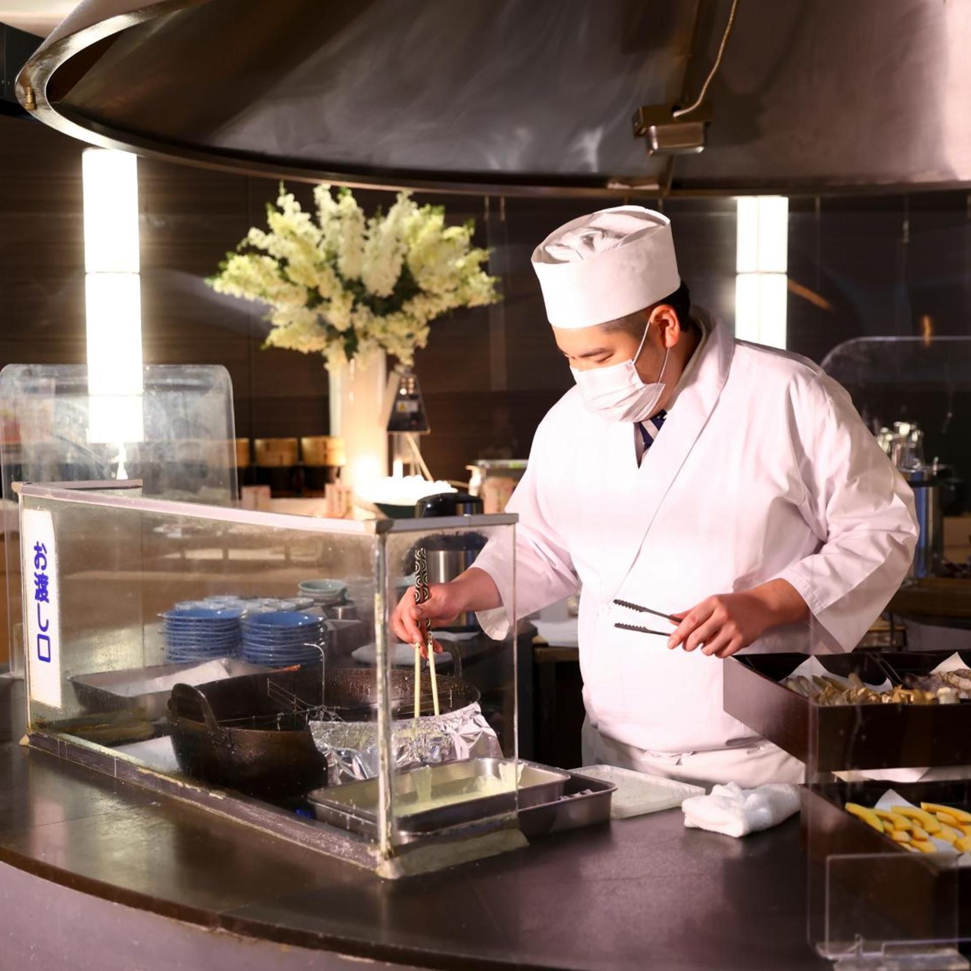 A chef in a white uniform preparing food in a commercial kitchen with stainless steel surfaces and a display case.