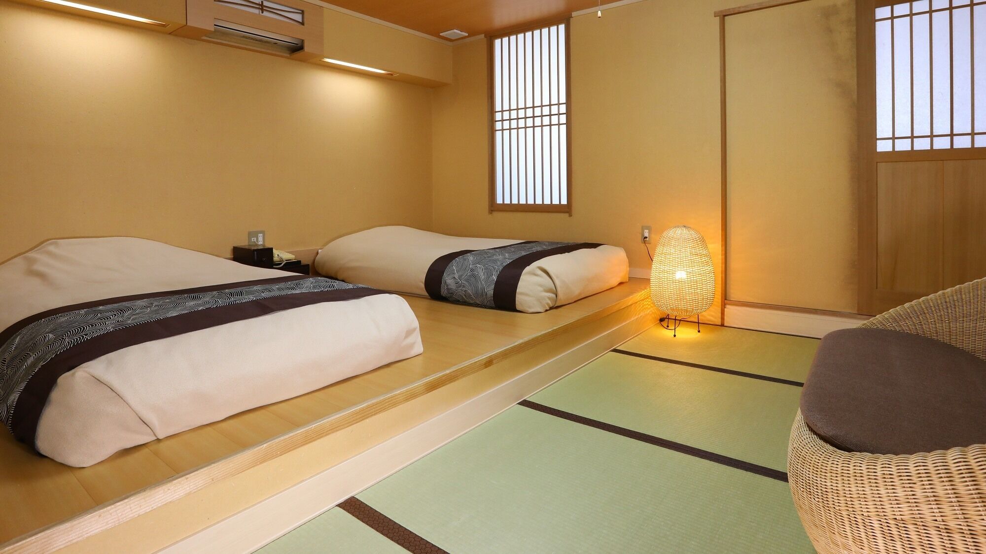 A traditional Japanese room with tatami flooring, two beds with futon-style bedding, a woven floor lamp, and sliding doors.