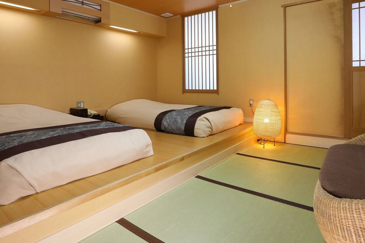 A traditional Japanese room with tatami flooring, two beds with futon-style bedding, a woven floor lamp, and sliding doors.