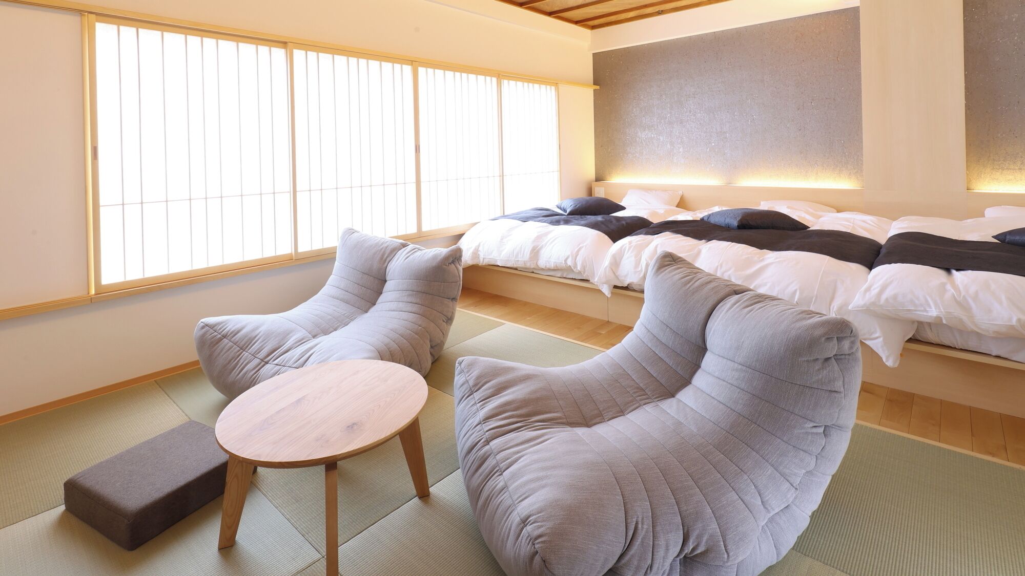 A room with tatami flooring, a bed with white bedding, and two grey armchairs with a small round table.