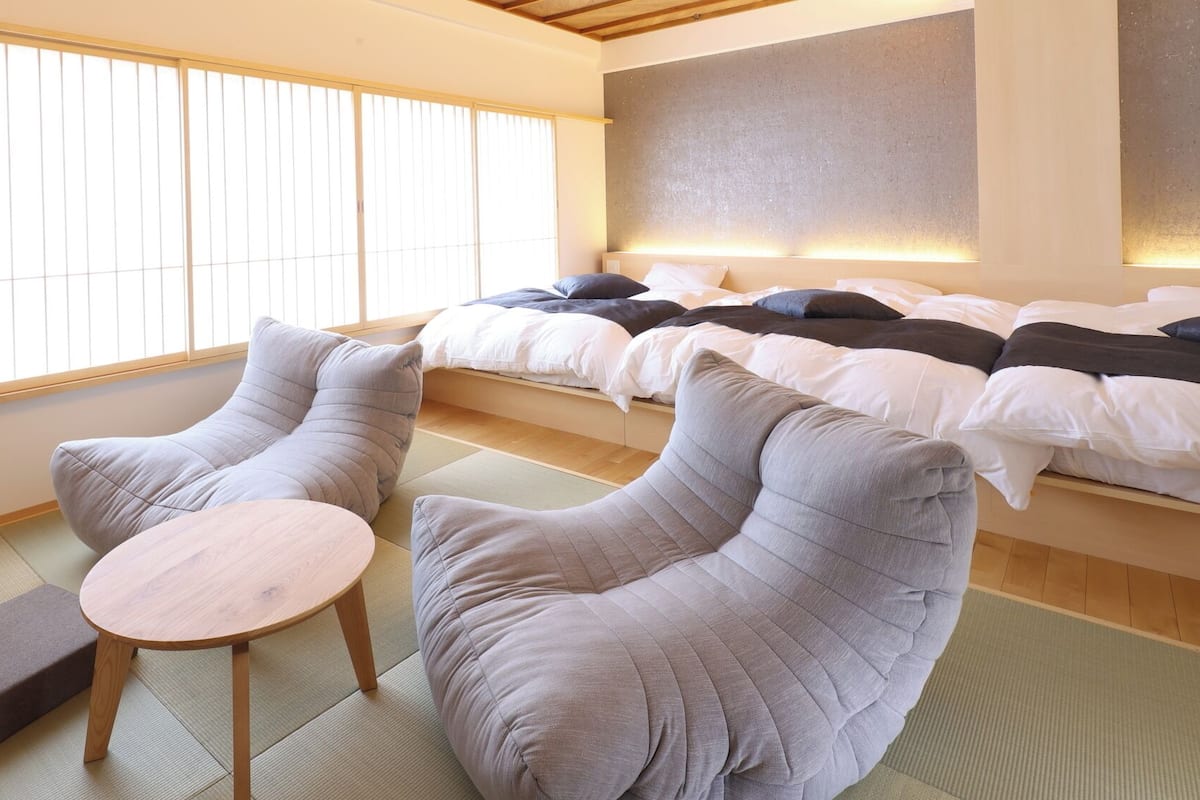 A room with tatami flooring, a bed with white bedding, and two grey armchairs with a small round table.