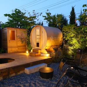 A wooden sauna structure with a door, a small wooden cabin, and a seating area with a bench and a chair.