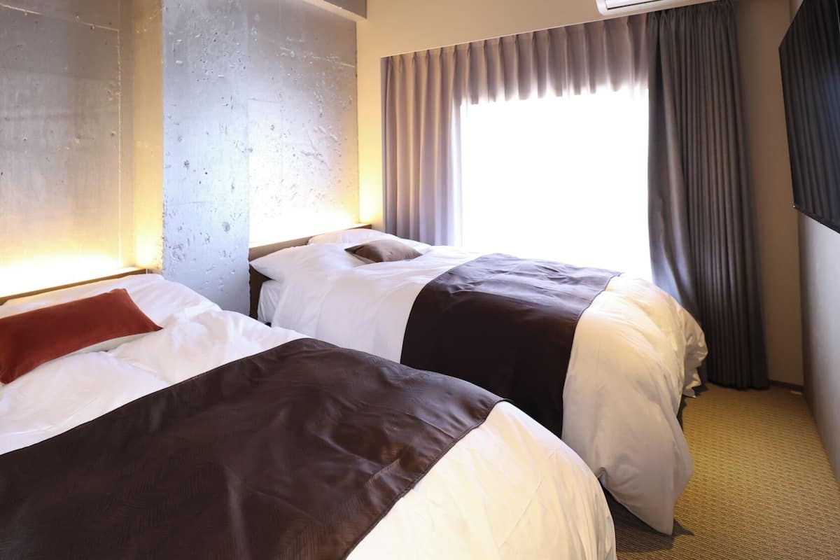 Two beds with white and brown bedding, a large window with curtains, and a wall with a textured surface.