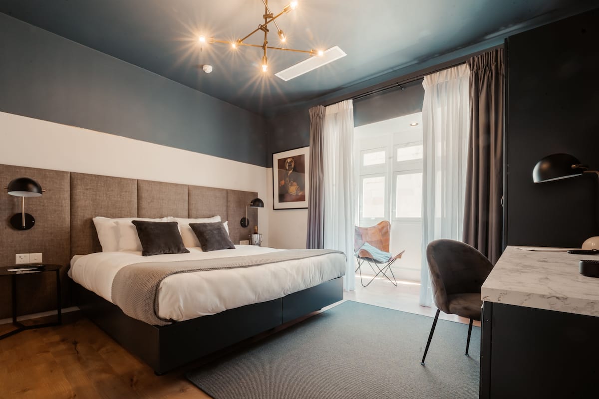 Une chambre moderne avec un grand lit, un bureau et une chaise. Il y a une fenêtre avec des rideaux et une vue sur l’extérieur.