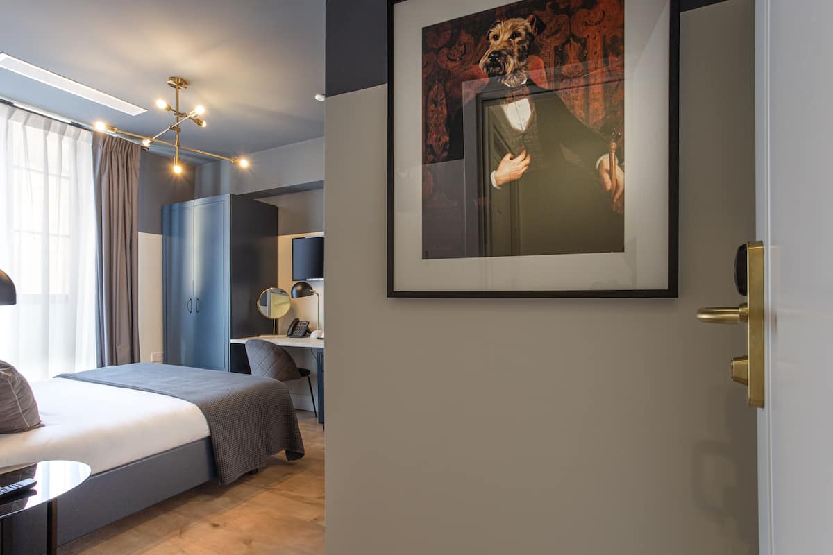 Une chambre d’hôtel moderne, équipée d’un lit, d’un bureau et d’une chaise, avec un tableau au mur et une porte dotée d’une poignée en laiton.