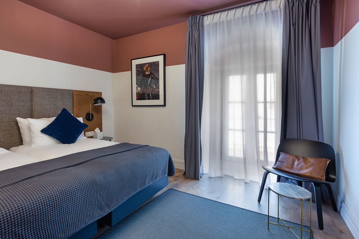 Une chambre d’hôtel avec un grand lit, une chaise, une petite table, un tableau au mur et une fenêtre avec des rideaux.