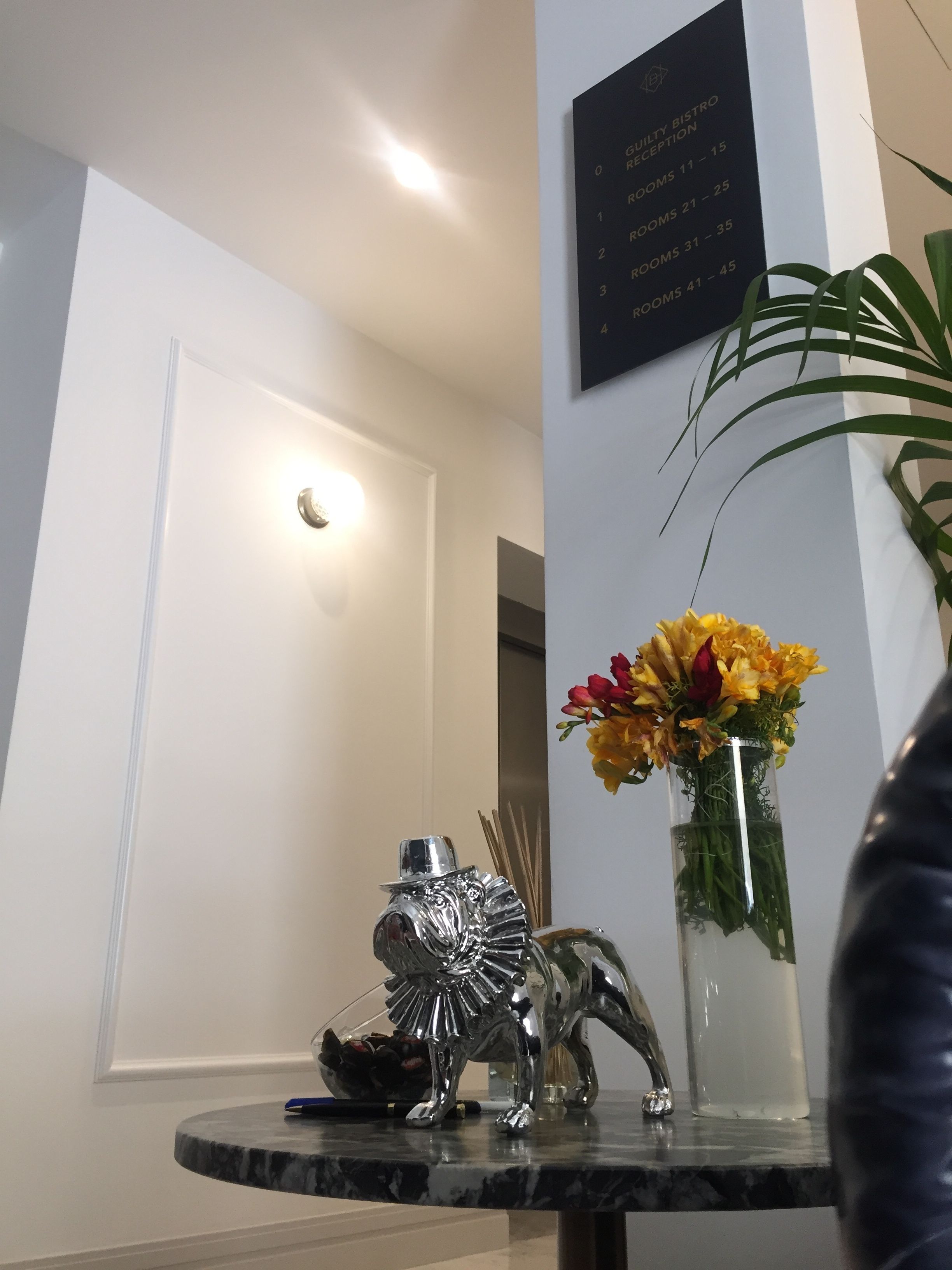 A reception area with a reception desk, a wall-mounted sign listing room numbers, a vase with flowers, and a decorative silver elephant sculpture.