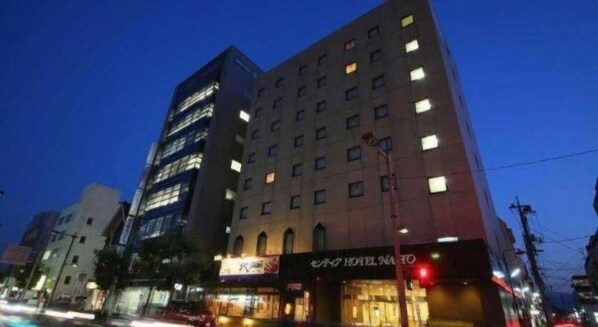 Un hôtel à plusieurs étages, avec des fenêtres illuminées et une enseigne indiquant « Hôtel Sano ».
