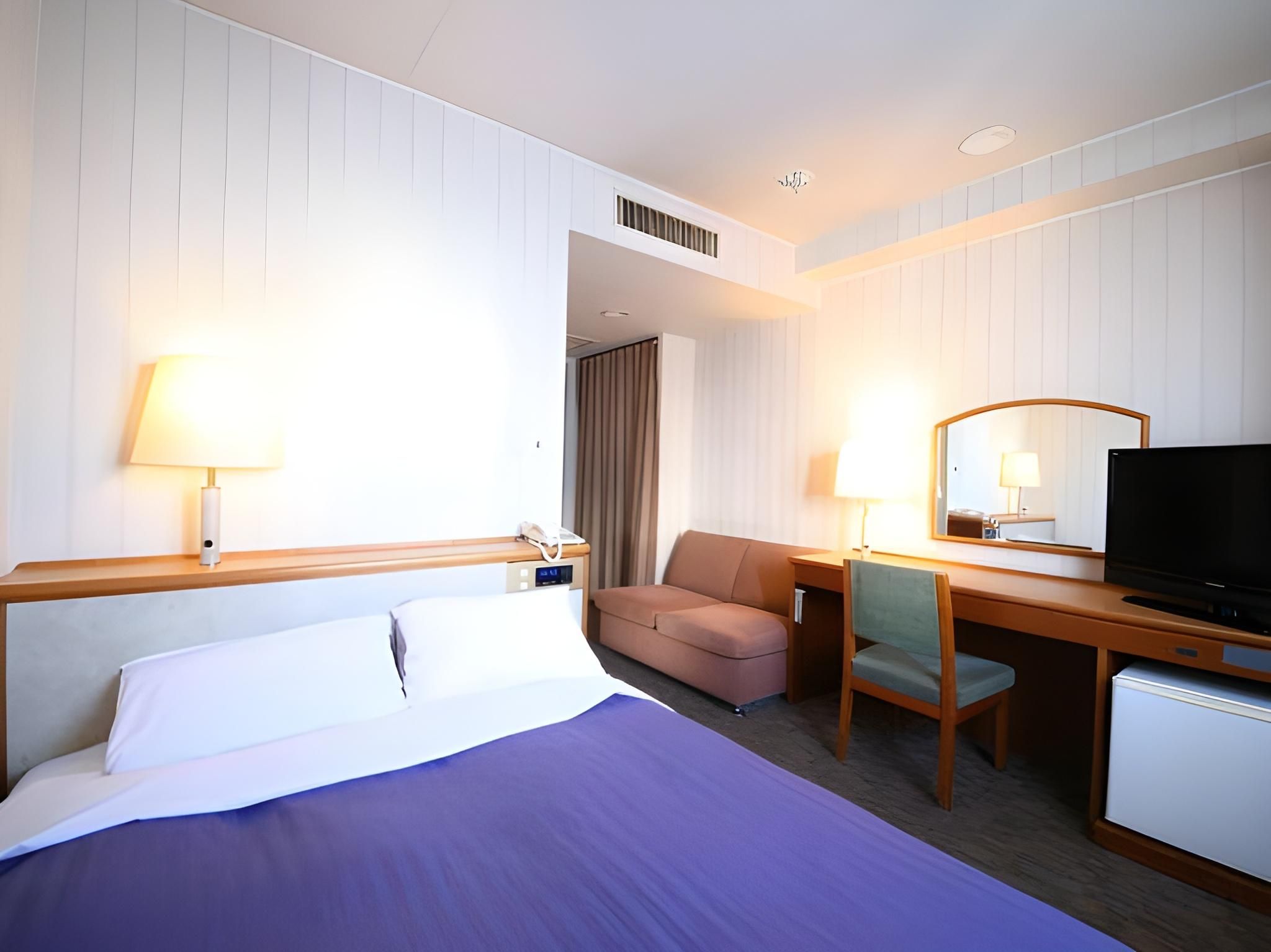 A hotel room with a bed, a desk, a chair, a television, and a small refrigerator.