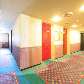 A hotel corridor with patterned carpet, red doors, and framed notices on the walls.