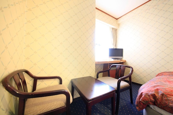 A hotel room with two wooden chairs, a small table, a TV on a stand, and a bed with a patterned blanket.