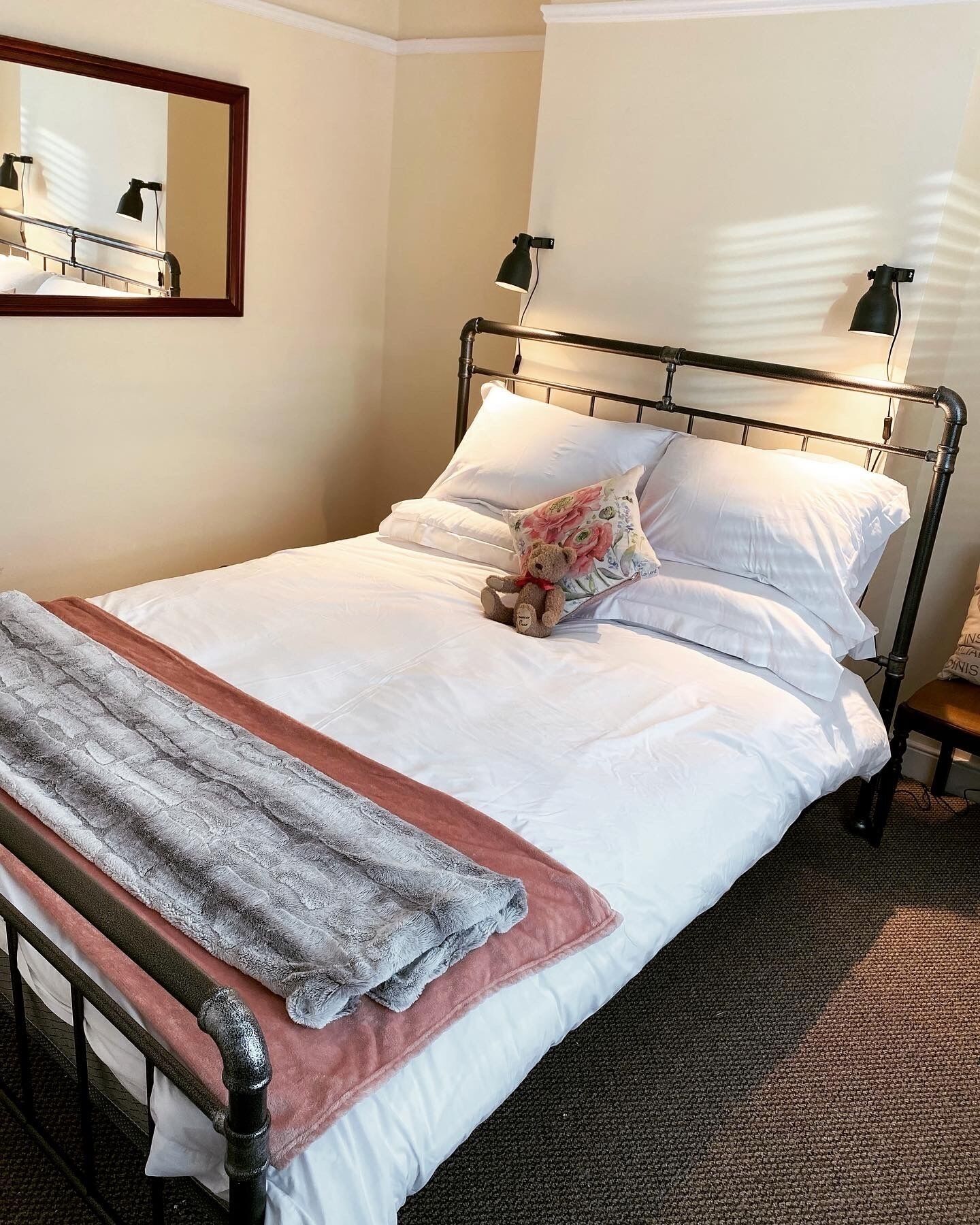 Een net opgemaakt bed met witte lakens, een grijze en roze deken en een teddybeer. Er staat een klein nachtkastje met een lamp en een flesje op.