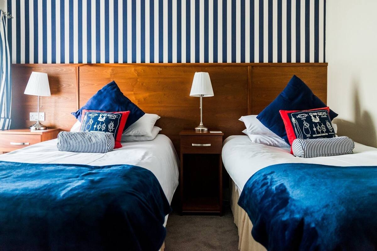 Ein Hotelzimmer mit zwei Betten, jeweils mit blauer Bettdecke und passenden Kissen. Es gibt zwei Nachttische mit Lampen und ein Holzkopfende.