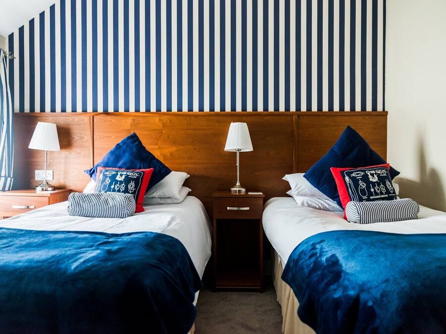 Une chambre d’hôtel avec deux lits, chacun recouvert d’une couverture bleue et agrémenté d’oreillers assortis. Il y a deux tables de chevet avec des lampes et une tête de lit en bois.