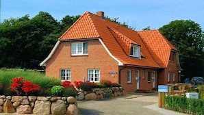 A two-story brick house with a red-tiled roof, a stone wall, and a well-kept garden with red flowers. - Apartment upper floor center (ID 184) (Sankt Peter-Ording)