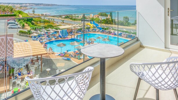 Un balcón con vistas a una piscina y una playa, con dos sillas blancas y una mesita.