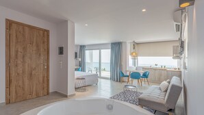 Una habitación de hotel moderna con una puerta grande de madera, una zona de estar con sofá y sillones, una mesa de comedor, un ventanal con vistas y un baño con bañera.