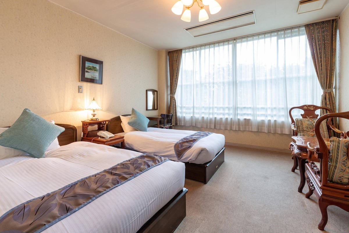 Une chambre d’hôtel avec deux lits, un bureau, une chaise, une lampe et une grande fenêtre avec des rideaux.