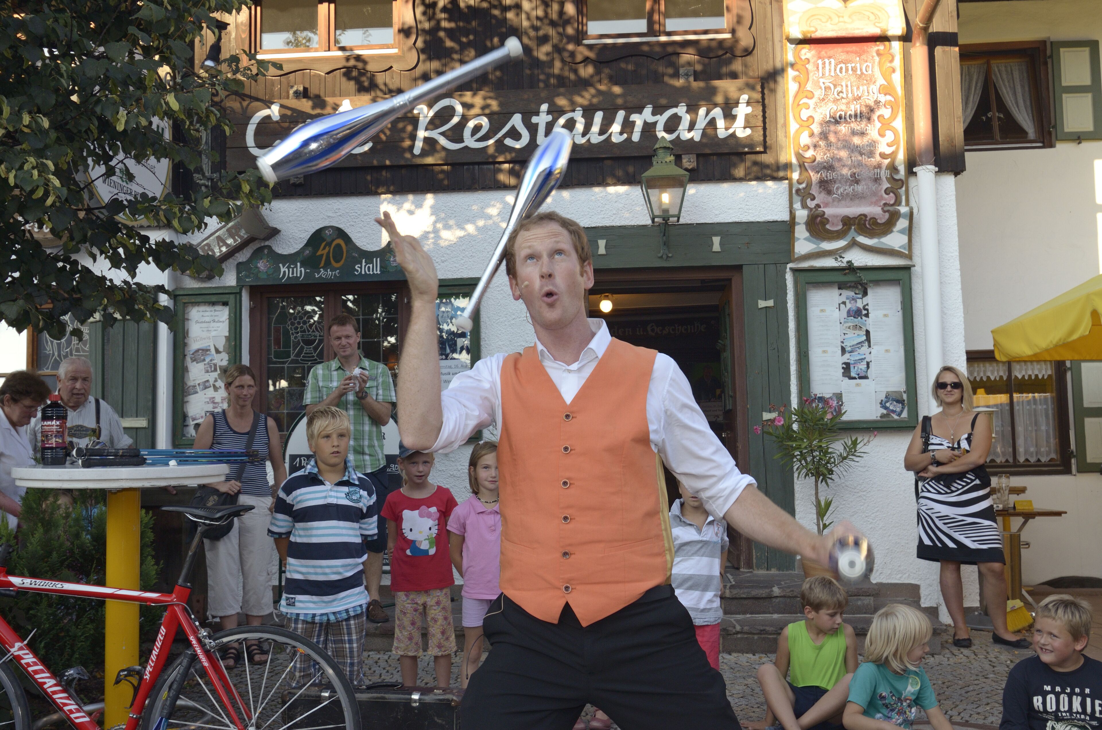 Eine Person jongliert vor einem Restaurant, dessen Schild 'Maria Halling Ladl' zeigt.
