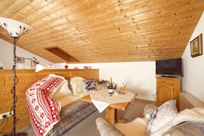 Ein gemütliches Zimmer mit Holzbalkendecke, einer Couch, einem Tisch mit Flasche und Gläsern, einem Fernseher und einem Schrank.