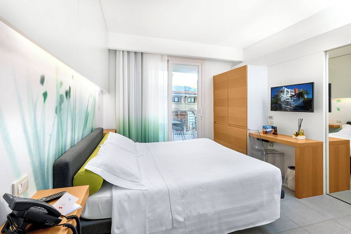 Habitación de hotel con cama, escritorio, silla, televisor y vista a edificios a través del ventanal.