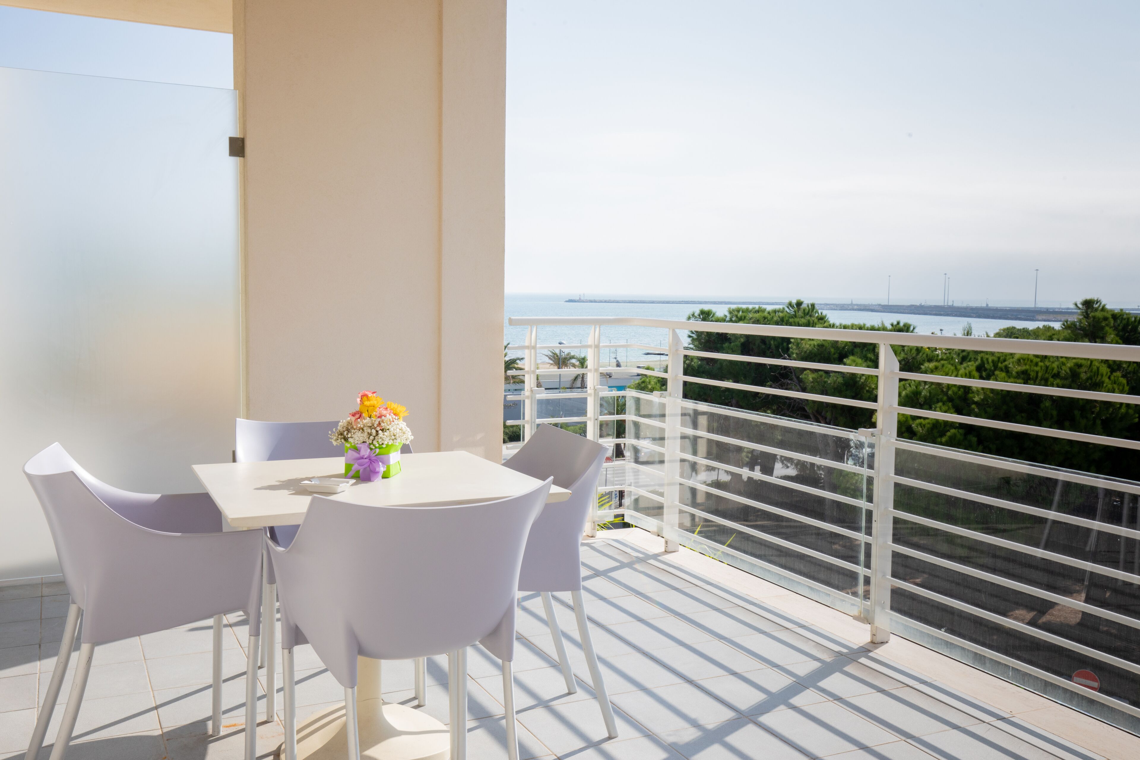 A balcony with a white table and chairs, a vase of flowers, and a view of the sea and distant buildings.