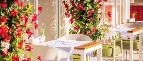 A dining area with tables and chairs, a potted plant with red flowers, and a window with a view.