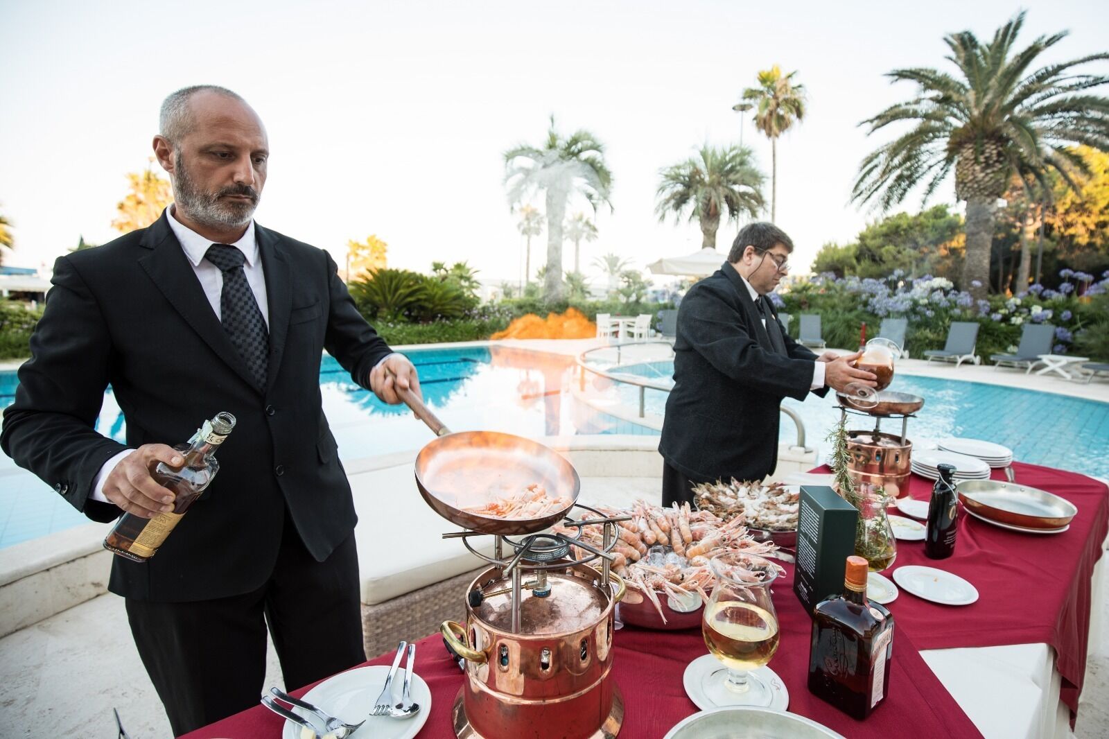 Tavolo apparecchiato per due in riva alla piscina, con una pentola di rame, una padella con cibo, una bottiglia di liquore e un bicchiere di vino.