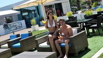 Un área junto a la piscina con sillones y mesas de mimbre, un letrero que dice 'Sabya Beach Garden' y un edificio con puertas de cristal al fondo.