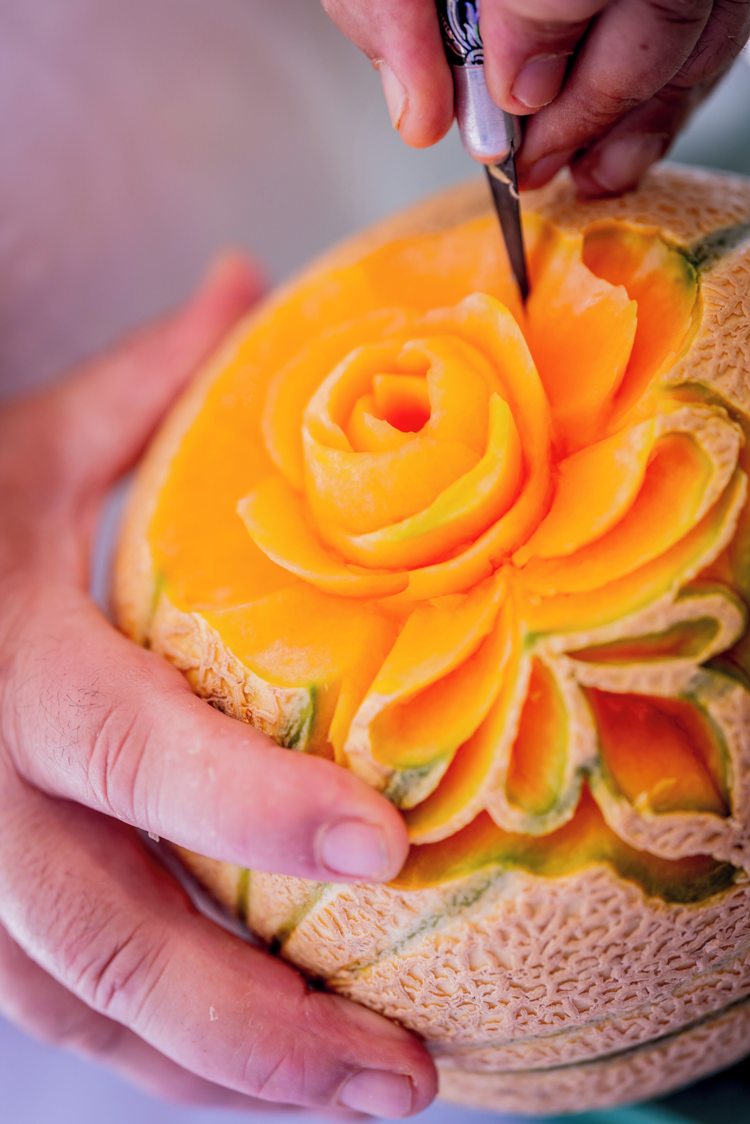 A person carving a detailed flower pattern into a cantaloupe.