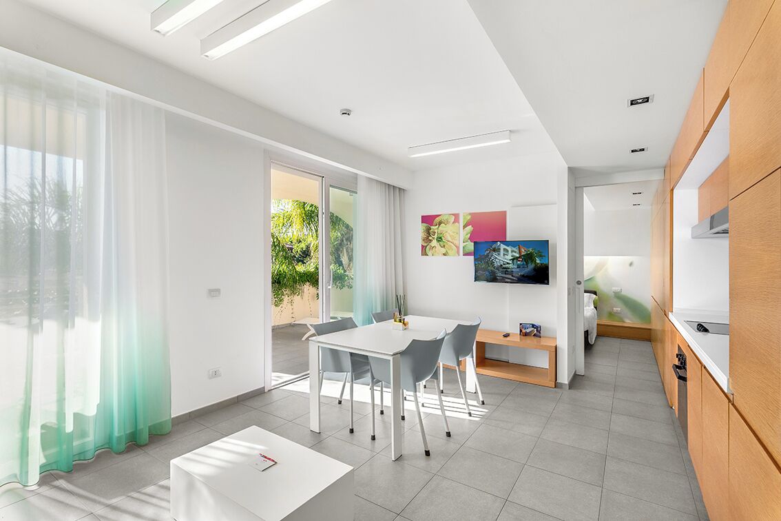 A modern kitchen with white cabinets, a dining area with a white table and chairs, and a TV mounted on the wall.